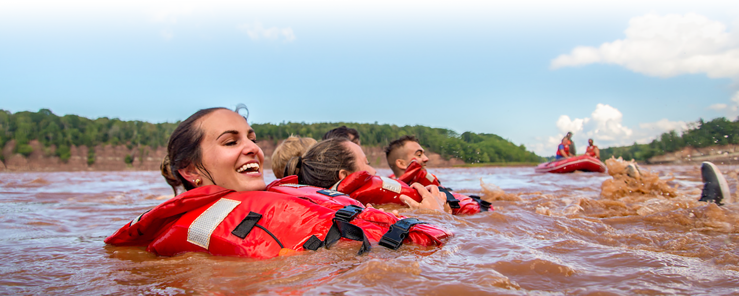 A group of young people wearing life jackets floating in the Shubenacadie River with a zodiac in the background.