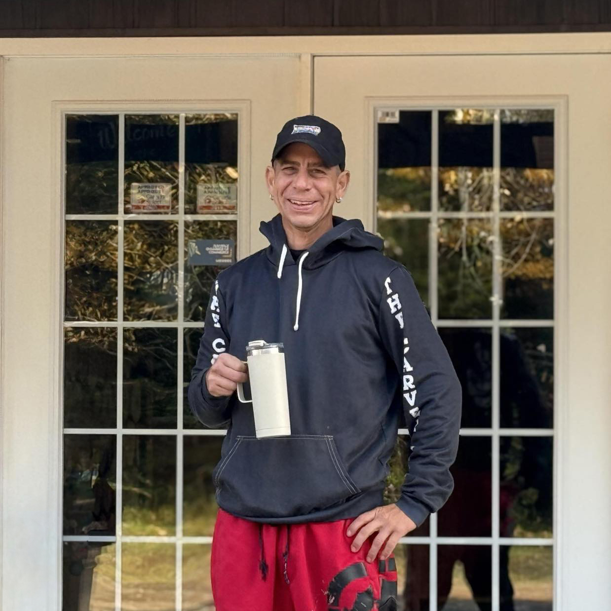 Jacob Paul, an employee of Tidal Bore Rafting Resort stands in front of the front entry way of the main lodge holding a coffee mug and smiling.