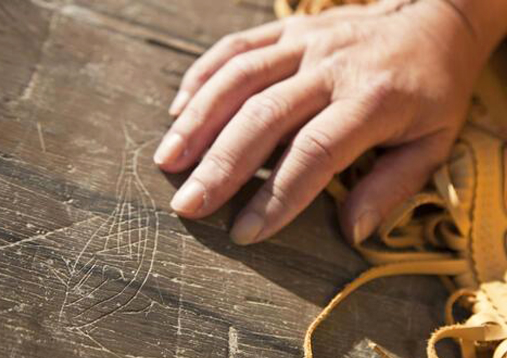 A hand pointing to a petroglyph of a whale carved into stone by Mi'kmaq people. The hand is resting on leather work.