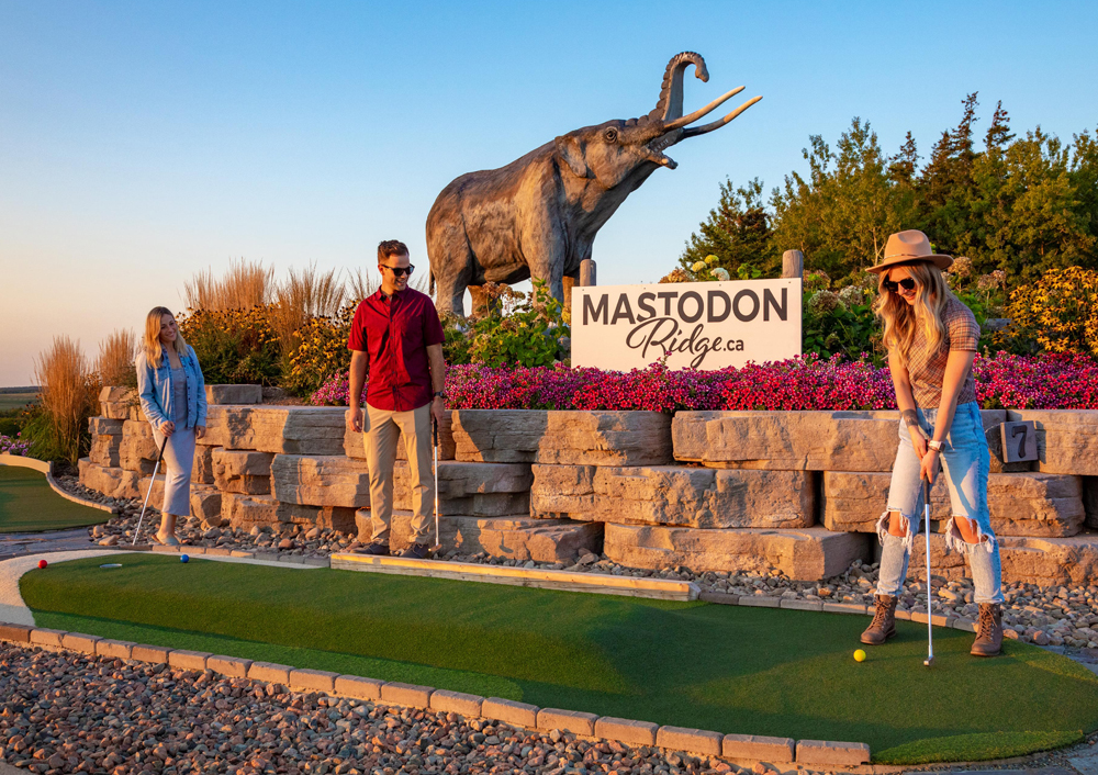 A young woman lines up a shot for mini golf in front of a large mastodon statue while two friends look on.