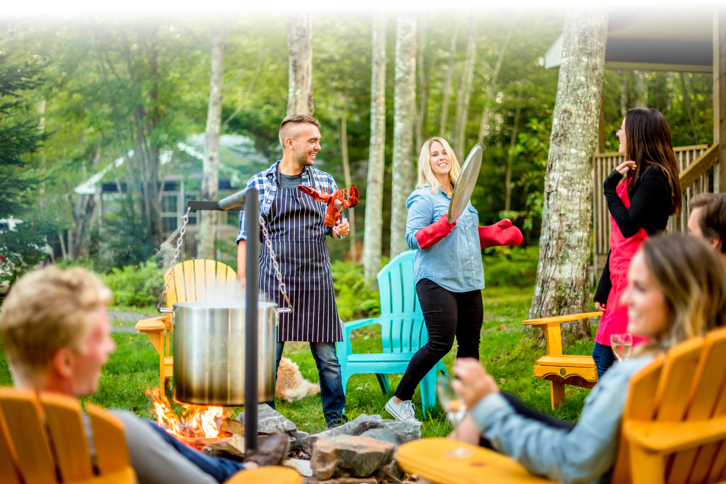 A man holds a lobster over a pot on an outdoor fire next to a woman wearing oven mitts.