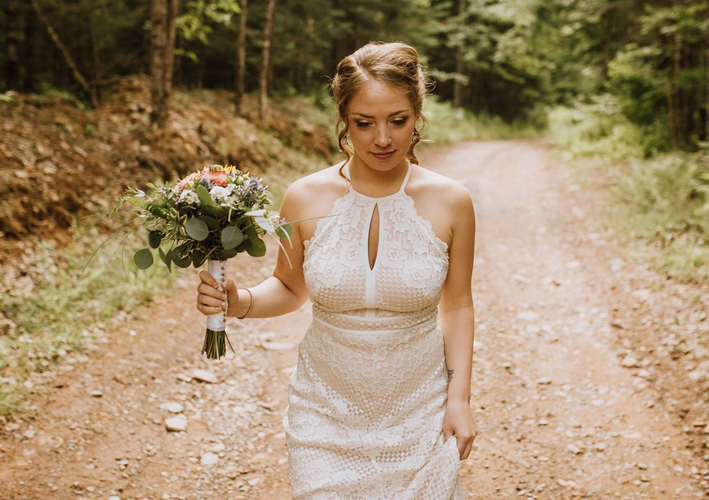 A woman wearing a wedding gown and holding a bouquet of flowers walks along a wooded trail at Tidal Bore Rafting Resort.